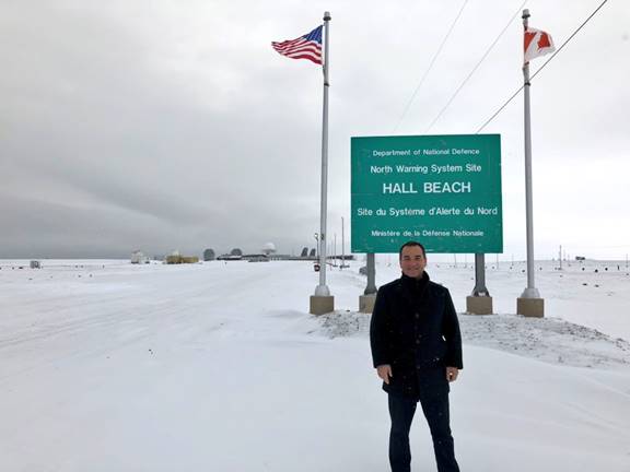 Photo du président du Comité des affaires étrangères se tenant dans la neige, à l’extérieur du site du système d’alerte du Nord, à Hall Beach. Il se trouve à côté de l’affiche du site, laquelle est flanquée d’un drapeau canadien et d’un drapeau américain.
