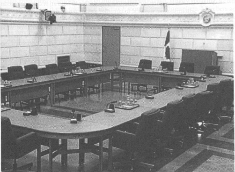 Photograph of a typical committee room, showing several tables placed in a rectangular format, surrounded by chairs. At the front of the room is a large podium for the console operator.